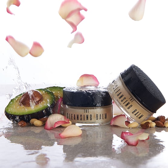 The Cleansing Balm by Sienna X. The image shows two jars of the product with rose petals and an avocado cut in half. These are some of the ingredients in the product.