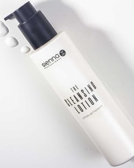 The cleansing lotion for the face by Sienna X. A white cylindrical bottle with a black pump action top is laid diagonally. Three pumps of the lotion are to the left of the pump action lid on the counter. All Sienna X packaging is recyclable.
