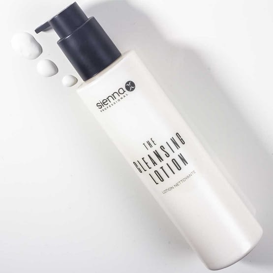 The cleansing lotion for the face by Sienna X. A white cylindrical bottle with a black pump action top is laid diagonally. Three pumps of the lotion are to the left of the pump action lid on the counter. All Sienna X packaging is recyclable.